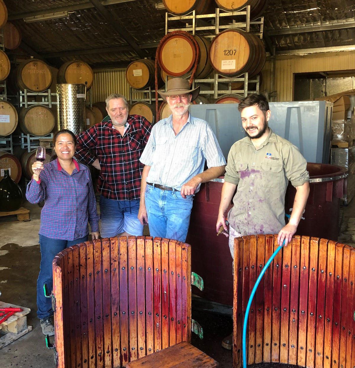 Four people stand in a winemaking cellar holding winemaking equipment and a wine glass