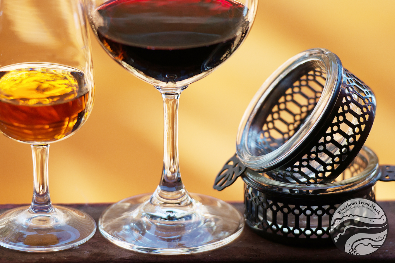 Artistically illuminated glasses of amber and red wine rest next to two decorative silver wine tasters